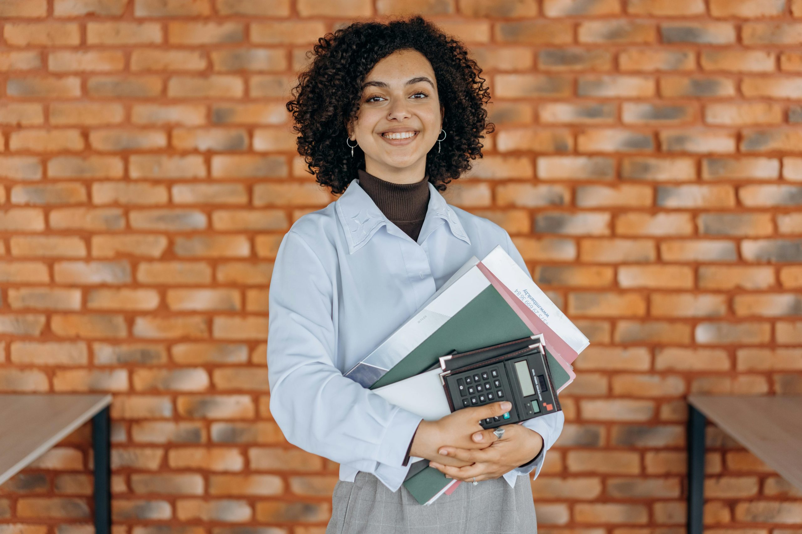 Cheerful woman with curly hair holding folders and a calculator against a brick wall.